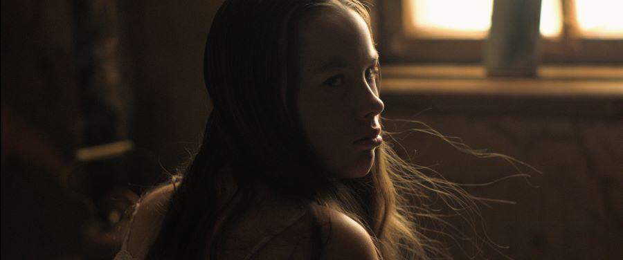 Ein junges Mädchen mit offenen langen Haaren sitzt vor einem Fenster und blickt über die rechte Schulter aus dem Augenwinkel auf 