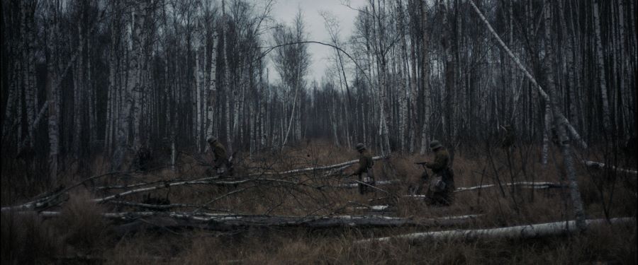 Drei Soldaten mit Gewehren in beigen Manteln bewegen sich in einem dichten brauen Herbstbrikenwald zu Fuß fort