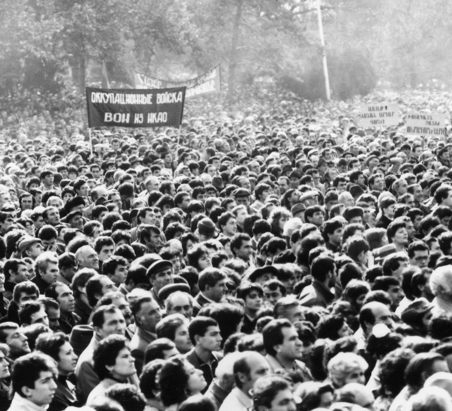 Bildunterschrift: Demonstration am Opernplatz, Erevan, November 1988, Die Übersetzung der Plakate der Demonstrant*innen groß in der Mitte: "Besatzungstruppen raus aus NKAO"; dahinter: "Armenier, lasst uns die Armenier in Artsakh unterstützen"; Rechts am Rand: "Nach vorne! Für eine gerechte Sache" und "Härteste Strafen für die Verbrecher".