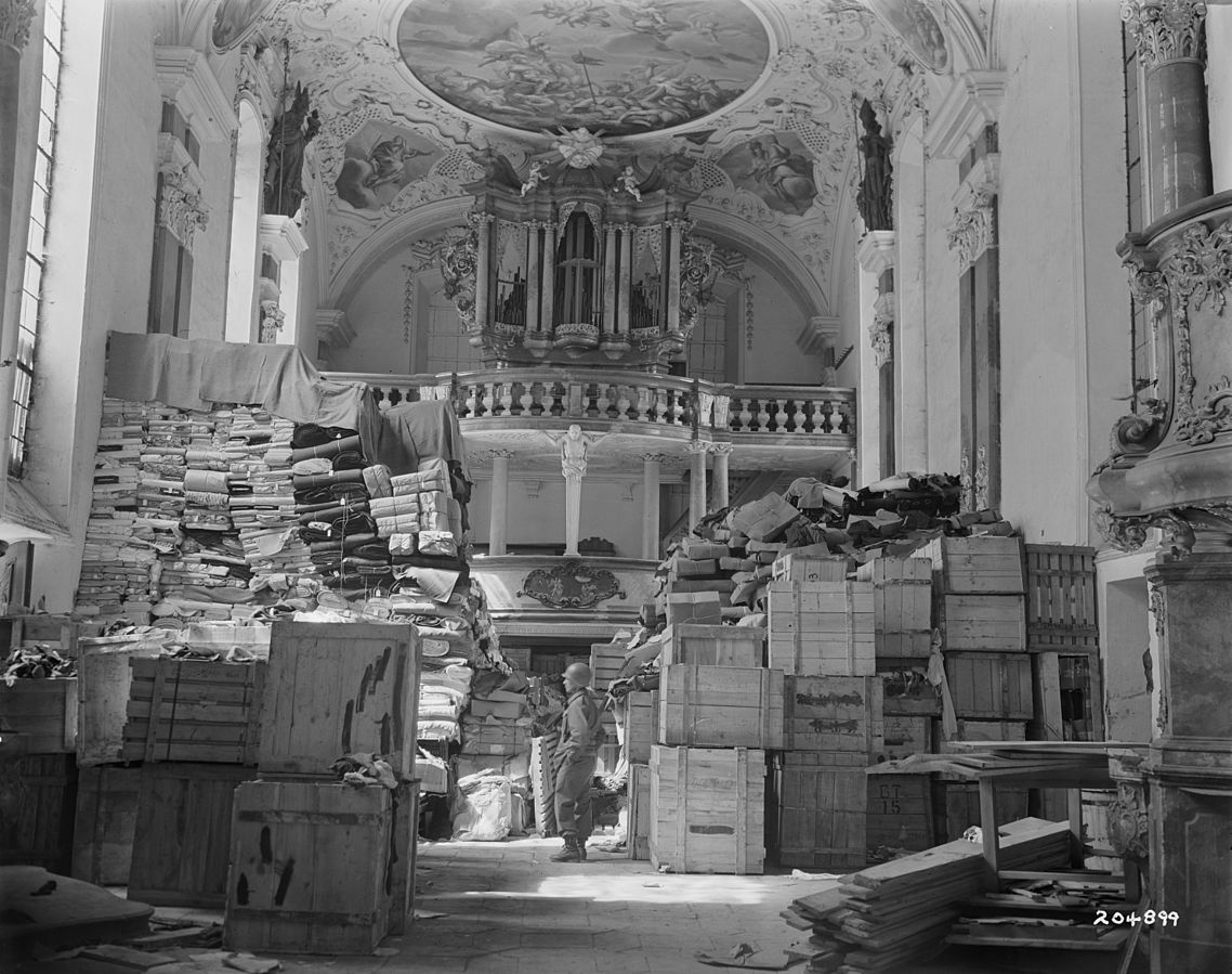Ein US-Soldat inmitten von aufgefundenen Kunst- und Kulturgütern in der Schlosskirche Ellingen (Bayern), Ende April 1945 // Nur ein kleiner Eindruck von den Dimensionen des nationalsozialistischen Kunstraubs: ein US-amerikanischer Soldat inmitten von aufgefundenen Kunst- und Kulturgütern in der Schlosskirche Ellingen (Bayern), Ende April 1945, Wikimedia Commons, Gemeinfrei.