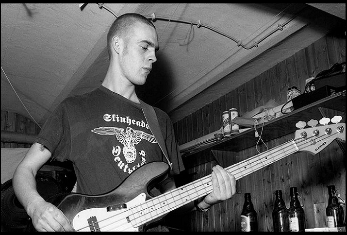 Mitglied einer Naziband spielt Gitarre und trägt ein T-Shirt mit Reichsadler und Aufschrift "Skinheads Deutschland", schwarzweiß