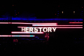 Das Wort "Herstory" in Leuchtbuchstaben auf einer verdunkelten Bühne.