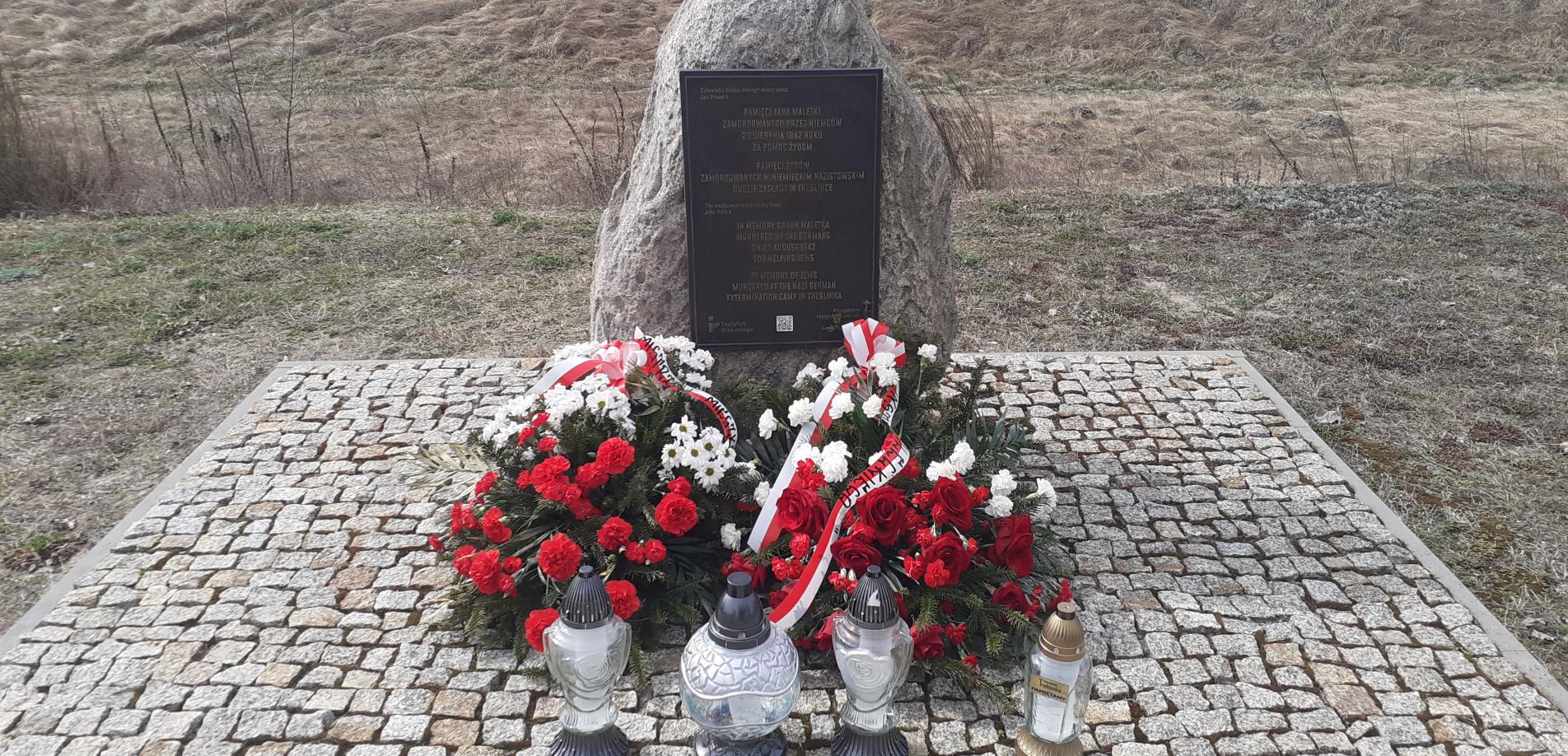 Denkmal an dem Ort der ehemaligen Bahnhofsstation im Dorf Treblinka, 31. März 2026. Die Kränze wurden vom Pilecki Institut (Witold Pilecki Institute of Solidarity and Courage) und vom Bürgermeister und den Bewohnern der Gemeinde Małkinia gestiftet; rechts eine goldene Kerze mit der goldenen Inschrift „Treblinka Museum – wir erinnern” 