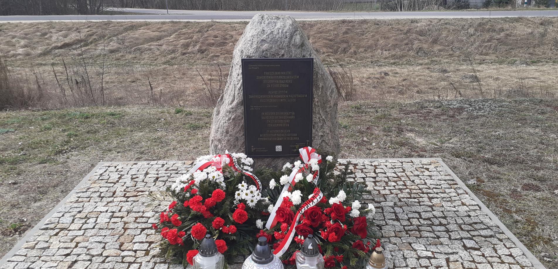 Denkmal an dem Ort der ehemaligen Bahnhofsstation im Dorf Treblinka, 31. März 2026. Die Kränze wurden vom Pilecki Institut (Witold Pilecki Institute of Solidarity and Courage) und vom Bürgermeister und den Bewohnern der Gemeinde Małkinia gestiftet; rechts eine goldene Kerze mit der goldenen Inschrift „Treblinka Museum – wir erinnern” 