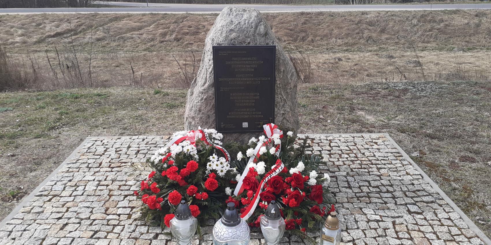Denkmal an dem Ort der ehemaligen Bahnhofsstation im Dorf Treblinka, 31. März 2026. Die Kränze wurden vom Pilecki Institut (Witold Pilecki Institute of Solidarity and Courage) und vom Bürgermeister und den Bewohnern der Gemeinde Małkinia gestiftet; rechts eine goldene Kerze mit der goldenen Inschrift „Treblinka Museum – wir erinnern” 