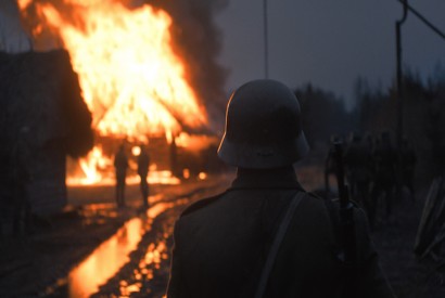 Eine Gruppe Soldaten und ein Soldat in Nahaufnahme von hinten in einer grauen Nacht stehen vor einem lichterloh brennenden Haus. Das Feuer reflektiert sich in den Pfützen